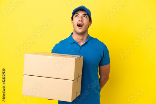 Delivery man over isolated yellow wall looking up and with surprised expression