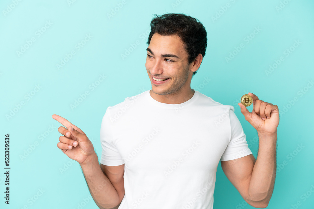 Young man holding a Bitcoin over isolated blue background pointing finger to the side and presenting a product Young man holding a Bitcoin over isolated blue background pointing finger to the side and presenting a product