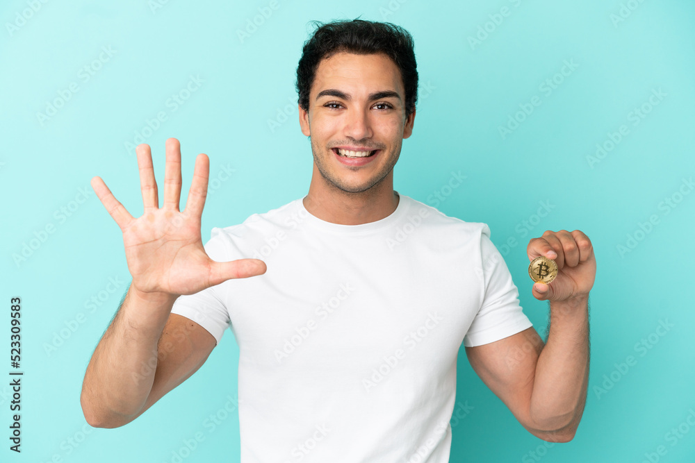 Young man holding a Bitcoin over isolated blue background counting five with fingers Young man holding a Bitcoin over isolated blue background counting five with fingers