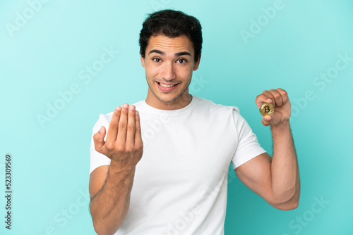 Young man holding a Bitcoin over isolated blue background inviting to come with hand. Happy that you came