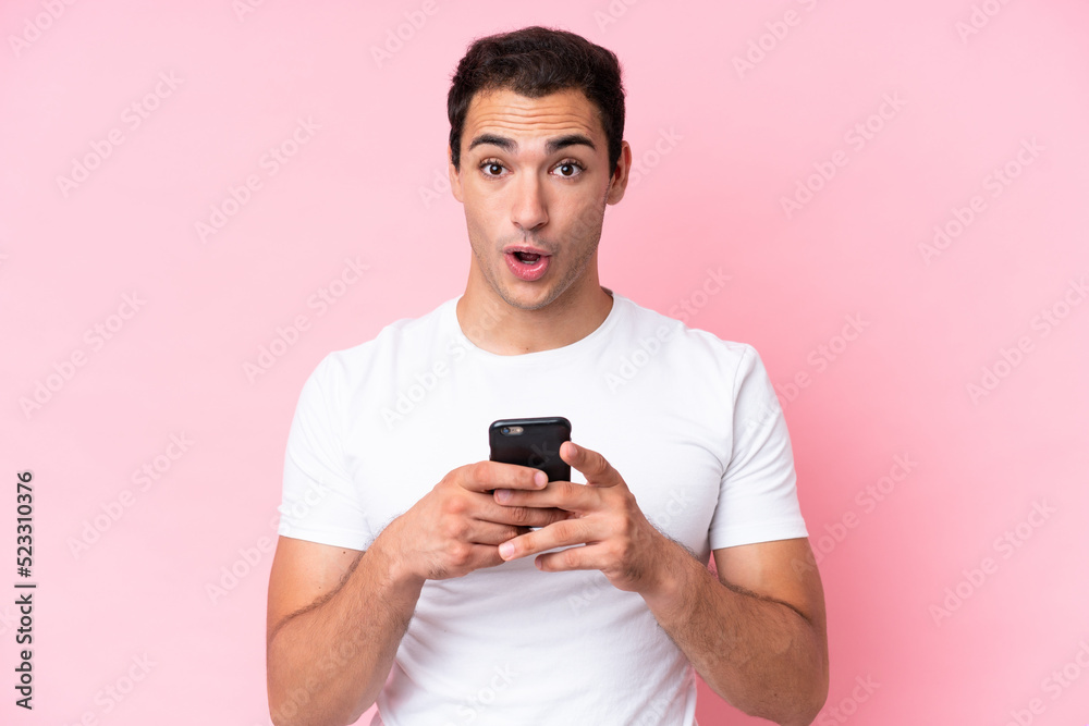 Young caucasian man isolated on pink background looking at the camera while using the mobile with surprised expression