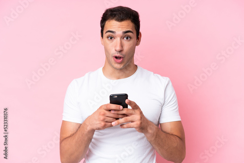 Young caucasian man isolated on pink background looking at the camera while using the mobile with surprised expression © luismolinero