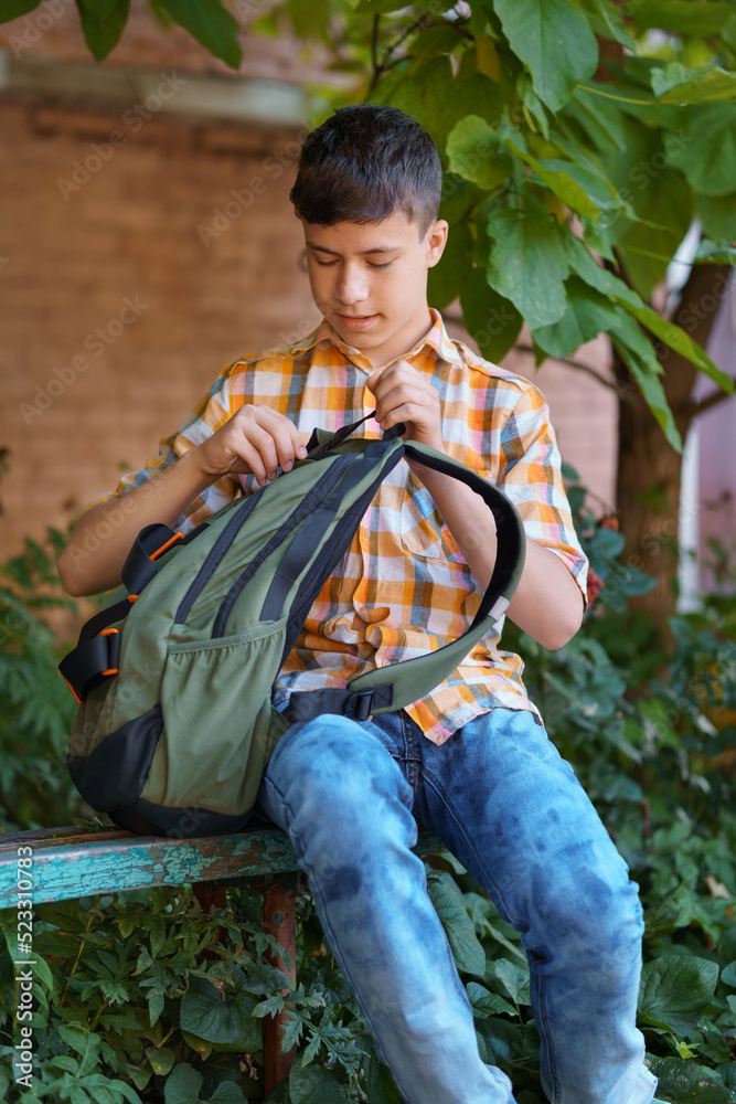 a teenage boy, being a student, is sitting on a bench near the school building, he takes a book or other school supplies out of his backpack