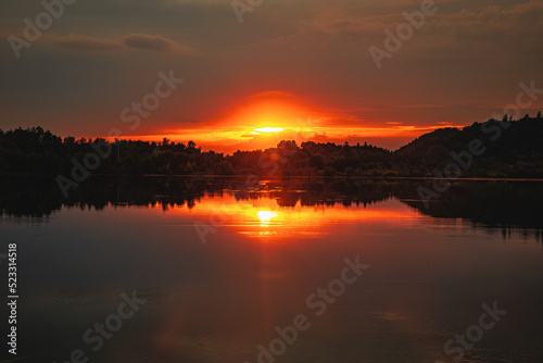 Epic red and gold clouds over forest lake at sunset. Dramatic cloud cover. Symmetrical reflections on the water, natural mirror. idyllic landscape.