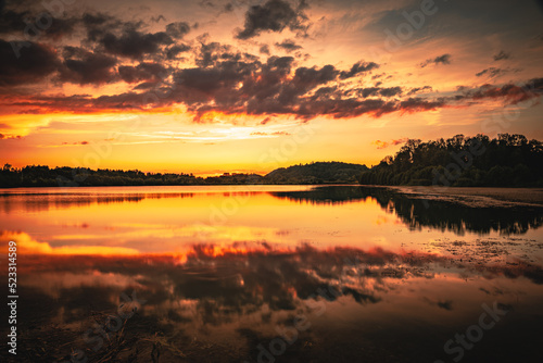 Epic red and gold clouds over forest lake at sunset. Dramatic cloud cover. Symmetrical reflections on the water, natural mirror. idyllic landscape. © CreativeImage