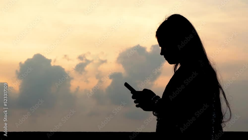 Silhouette of a cheerful attractive young Indian woman holding cell phone and smiling, browsing internet or texting concept isolated outdoor background