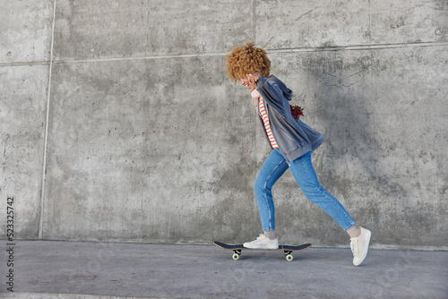 Energetic hipster girl rides skateboard dressed in casual clothes enjoys spare time poses against grey concrete wall with blank space for your promotional content. People pastime and recreation