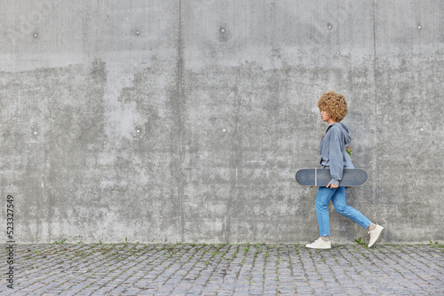 Active teenage girl with curly hair dressed in hoodie jeans and sneakers carries longboard enjoys active rest strolls outdoors against grey concrete wall empty space for your promotional content