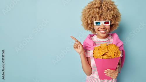 Happy curly haired young woman invites you to cinema points away on blank space advertises intersting film to watch holds paper bucket of crisps wears 3d glasses poses against blue background. photo