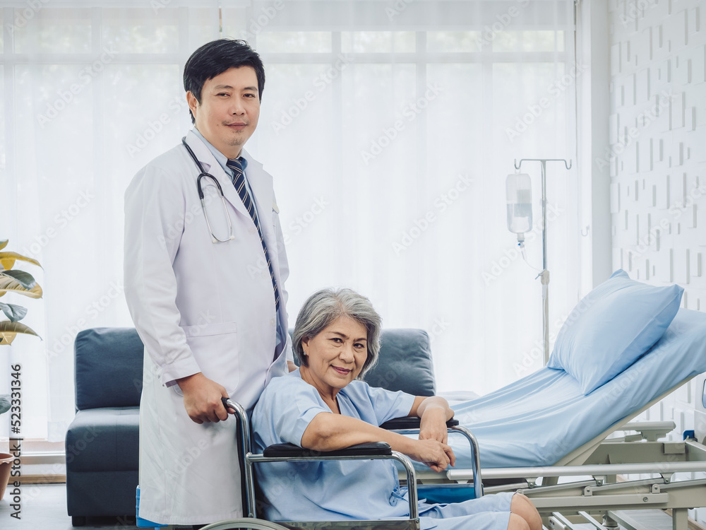 Asian elderly female patient dressed in light blue, smiling happily sits in wheelchair with kindly male doctor in white suit standing and carrying the wheelchair near bed in recovery room in hospital. Asian elderly female patient dressed in light blue, smiling happily sits in wheelchair with kindly male doctor in white suit standing and carrying the wheelchair near bed in recovery room in hospital.