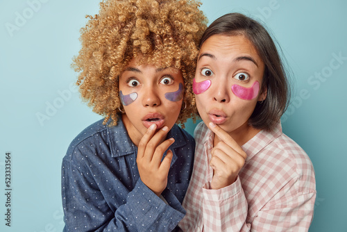 Amazed diverse female models stare stupefied apply beauty patches under eyes for skin treatment hold breath from astonishment dressed in nightwear isolated over blue background. Wellness concept