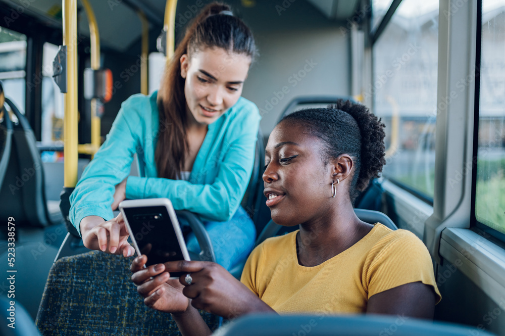 Multiracial friends talking and using a smartphone while riding a bus in the city Multiracial friends talking and using a smartphone while riding a bus in the city