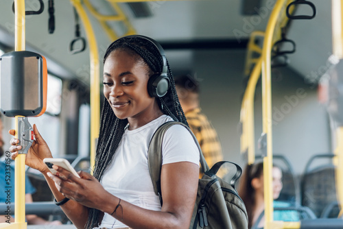 Fototapeta African american woman riding a bus and using a smartphone and headphones