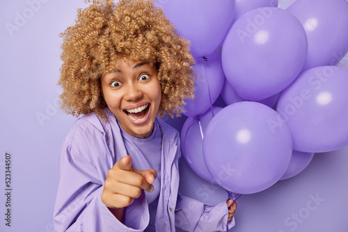 Positive surprised woman with curly hair points index finger forward smiles broadly wears jacket shows something impressive holds bunch of inflated helium balloons isolated over purple background