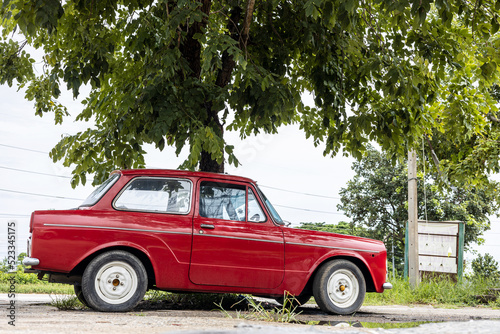A view of the old classic red sedan parked in the rain for a long time under the leafy green trees.