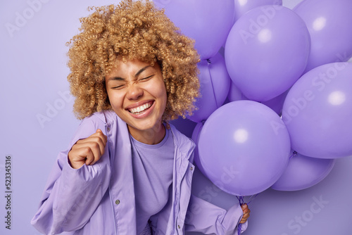 Happy carefree woman with curly blonde hair grins at camera keeps eyes closed dressed in stylish jacket celebrates special occasion holds bunch of inflated helium balloons isolated over purple wall