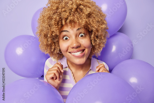 Surprised curly haired woman bites lips looks wondered dressed in jumper poses around inflated balloons celebrates special occasion isolated over purple background. People and holiday concept
