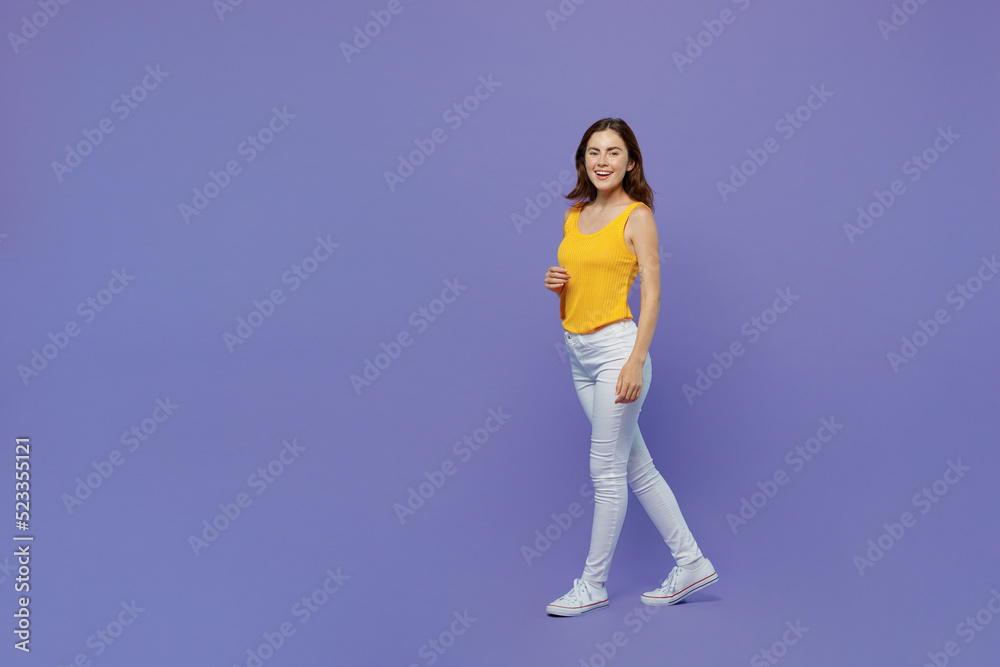 Full body side view young happy fun woman 20s she wear yellow tank shirt look camera walking going strolling isolated on plain pastel light purple background studio portrait. People lifestyle concept.