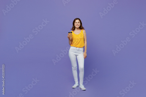 Full size young woman 20s she wear yellow tank shirt hold takeaway delivery craft paper brown cup coffee to go isolated on plain pastel light purple background studio portrait People lifestyle concept