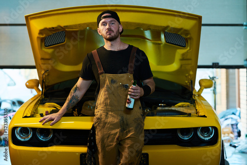 Young male technician or engineer of maintenance service with bottle of beer standing against yellow car with open hood in workshop