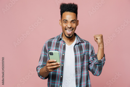 Young smiling happy man of African American ethnicity 20s wear blue shirt hold in hand use mobile cell phone do winner gesture isolated on plain pastel light pink background. People lifestyle concept.