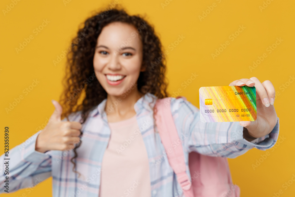 Young happy black teen girl student she wear casual clothes backpack bag hold credit bank card show thumb up isolated on plain yellow background. High school university college concept. Focus on card. Young happy black teen girl student she wear casual clothes backpack bag hold credit bank card show thumb up isolated on plain yellow background. High school university college concept. Focus on card.