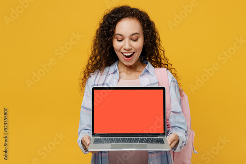 Young black teen girl student she wear casual clothes backpack bag hold laptop pc computer blank screen workspace area isolated on plain yellow color background High school university college concept.