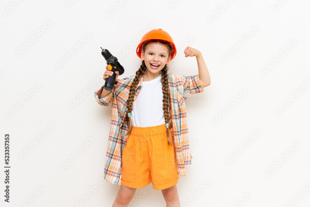 A child in a construction helmet and with a drill in his hands on a white isolated background. A little girl with long pigtails is getting ready for renovation in the nursery.