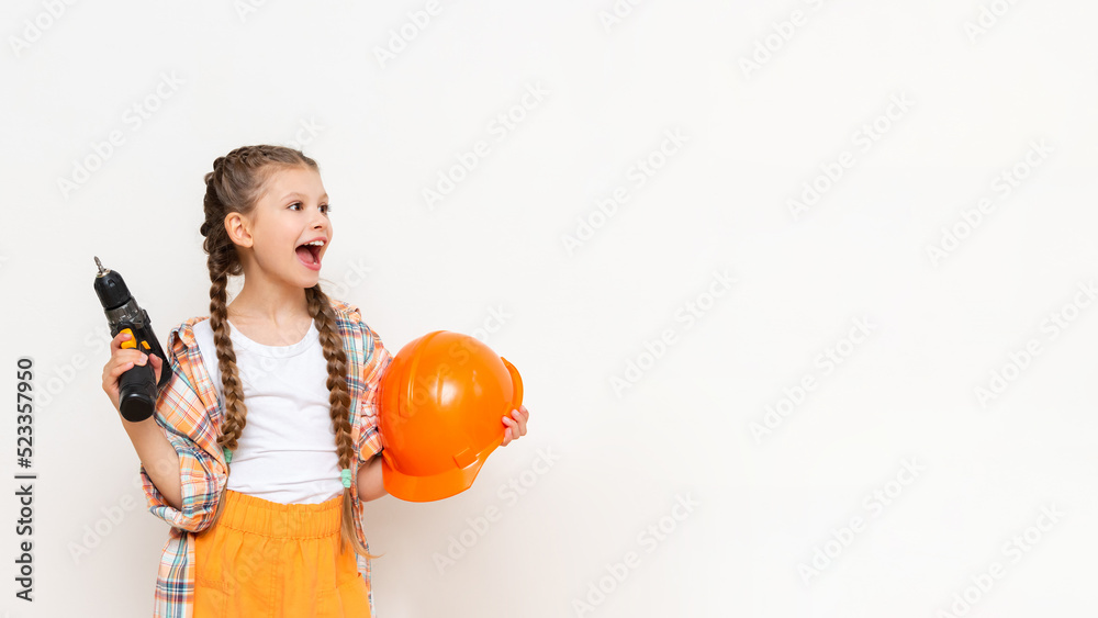 The child is holding a cordless drill and a protective construction helmet on a white insulated background. A little girl with long pigtails holding a screwdriver is smiling broadly. Copy Space. The child is holding a cordless drill and a protective construction helmet on a white insulated background. A little girl with long pigtails holding a screwdriver is smiling broadly. Copy Space.