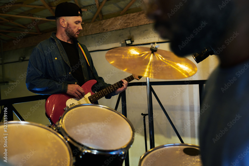 Obraz premium Young man in casualwear plucking strings of electric guitar during music repetition while standing in front of drum set and percussionist