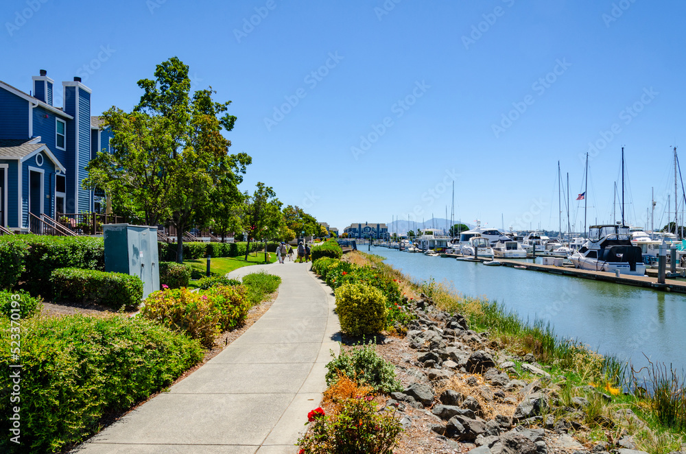 Footpath around the edge of Benicia Marina in California, USA past colourful blue houses and planting.