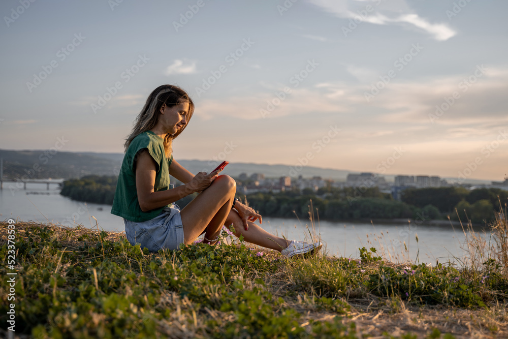 Young woman in green shirt sitting on grass with crossed legs. She is using her smartphone. Side photography. Cityscape in background.
