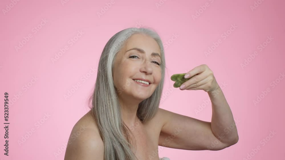 Beauty care procedures. Happy beautiful grey haired senior lady doing skin lifting massage with gouache stone Beauty care procedures. Happy beautiful grey haired senior lady doing skin lifting massage with gouache stone