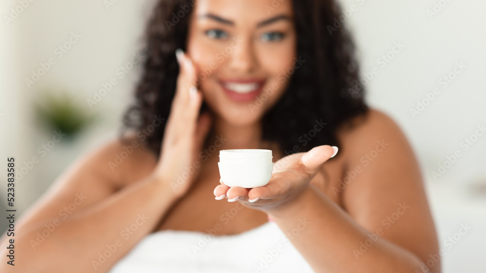 African american plus size woman holding jar of skin cream, applying moisturiser on face after shower, selective focus African american plus size woman holding jar of skin cream, applying moisturiser on face after shower, selective focus