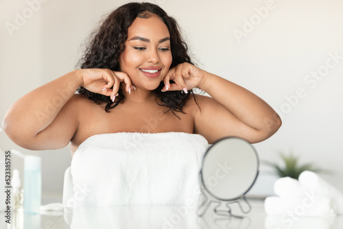 Beautiful black chubby lady making lymphatic drainage self massage, sculpting jaw line in front of mirror, free space