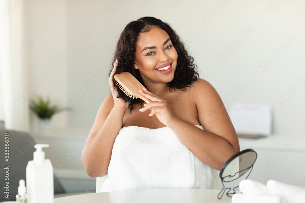 Happy black chubby woman brushing her curly hair with comb and making haircare routine at home Happy black chubby woman brushing her curly hair with comb and making haircare routine at home