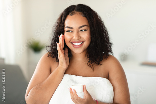 Happy black overweight lady using moisturising face cream, holding jar with beauty product and smiling