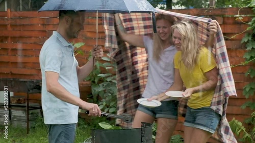 Picnic in nature. man grilling delicious meal in rain. Beautiful young girls cheerfully run up, covered with blanket from rain laugh and serve plate for fried meat. Family picnic on summer day outside