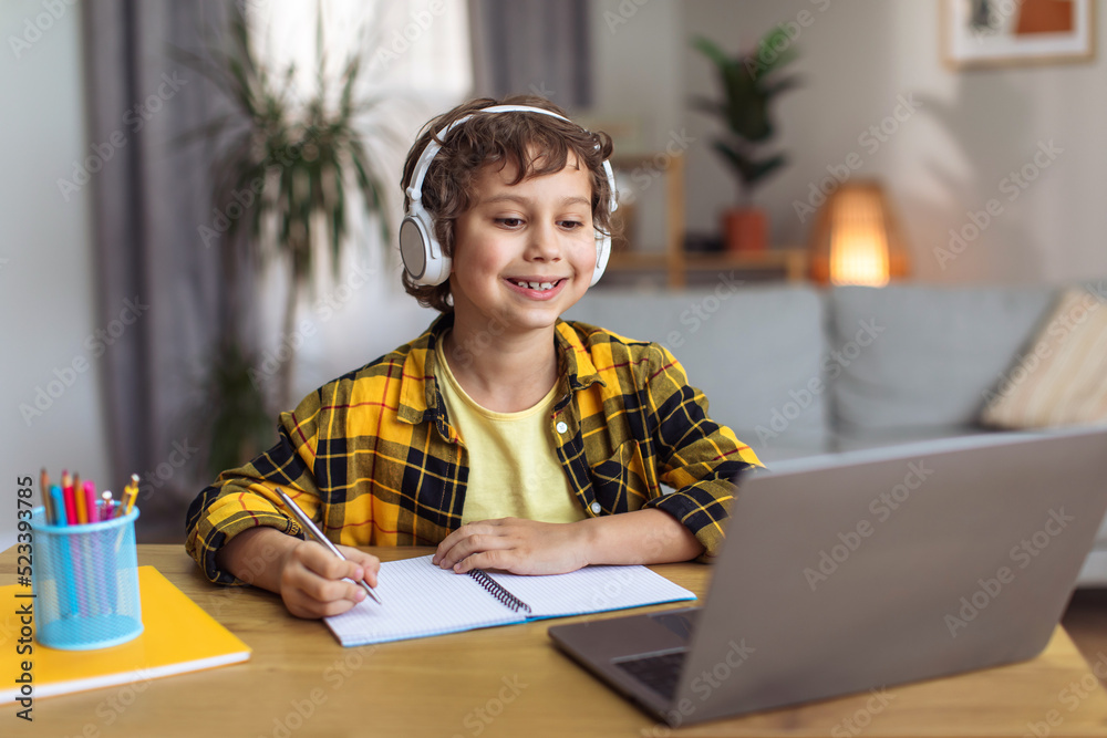 Cute happy little schoolboy enjoying video lesson at home, video chatting with teacher and writing notes, free space Cute happy little schoolboy enjoying video lesson at home, video chatting with teacher and writing notes, free space