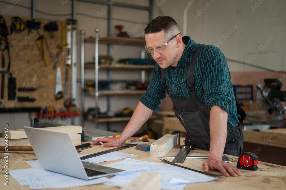 Portrait of a carpenter in protective glasses and work overalls uses a laptop in a workshop. Portrait of a carpenter in protective glasses and work overalls uses a laptop in a workshop.