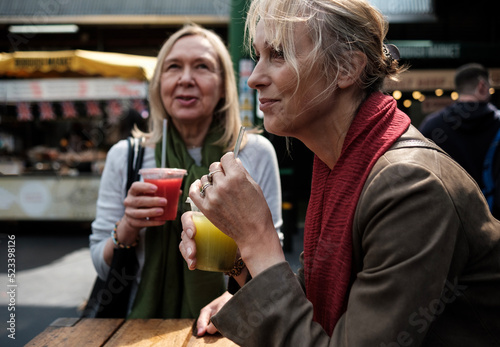 Portrait of two mature women friends sitting on a table in a street market drinking juices.