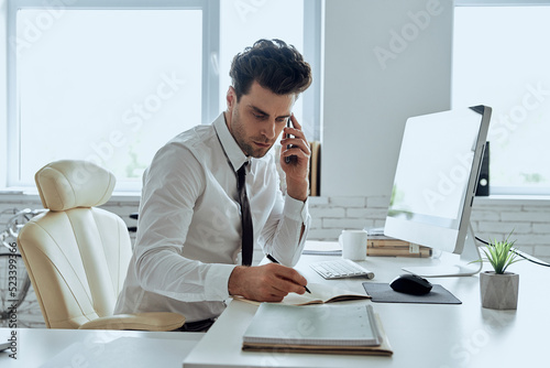 Confident man in shirt and tie making notes and talking on phone while sitting in office