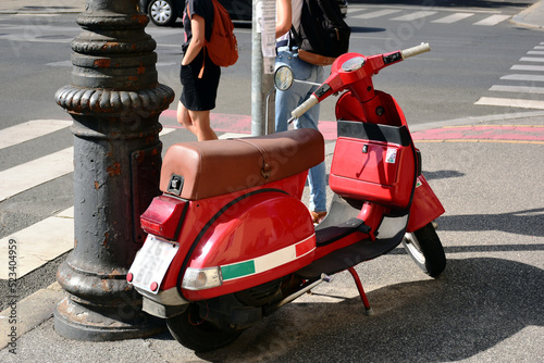Popular brand Italian made red, white and green scooter closeup at street corner. city and urban transportation concept. asphalt pavement. white pedestrian crossing. young woman and man