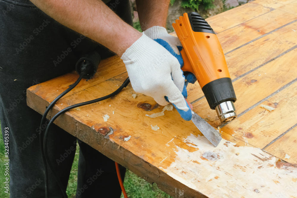 Man removing old varnish and paint from wood using scraper and heat gun. Restoration work of old wooden table outdoors. Furniture repair, reuse of old things concept. - obrazy, fototapety, plakaty 