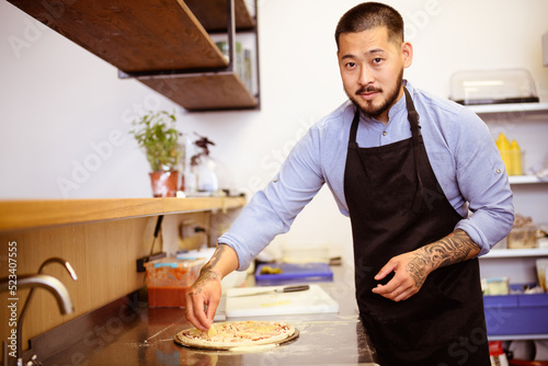 Asian chef putting pineapple on pizza and looking at camera in cafe kitchen 