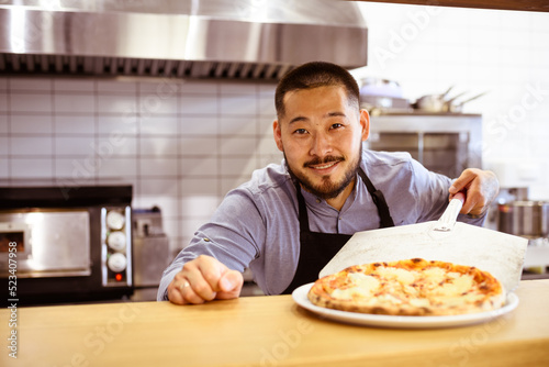 Smiling asian chef looking at camera near fresh pizza in cafe 