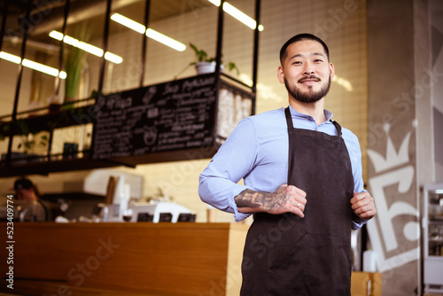 Tattooed asian small business owner in apron smiling at camera in cafe 