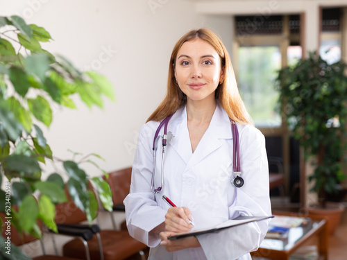 Portrait of smiling young woman doctor wearing white coat meeting patient in medical office, filling out medical form at clipboard