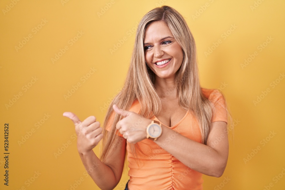 Young woman standing over yellow background pointing to the back behind with hand and thumbs up, smiling confident Young woman standing over yellow background pointing to the back behind with hand and thumbs up, smiling confident
