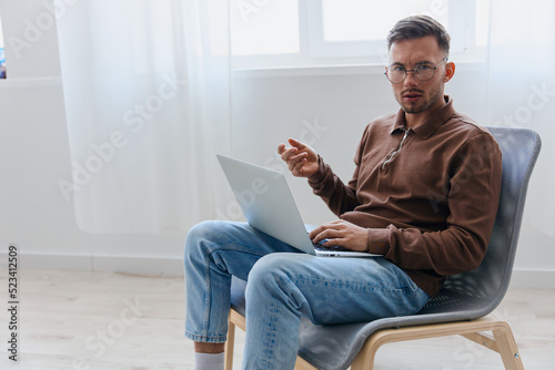 Pensive serious young man in eyewear raise hand up thinking about best solution for project looks at camera sitting in chair at home. Remote Job Distant Work Communication Modern Profession concept